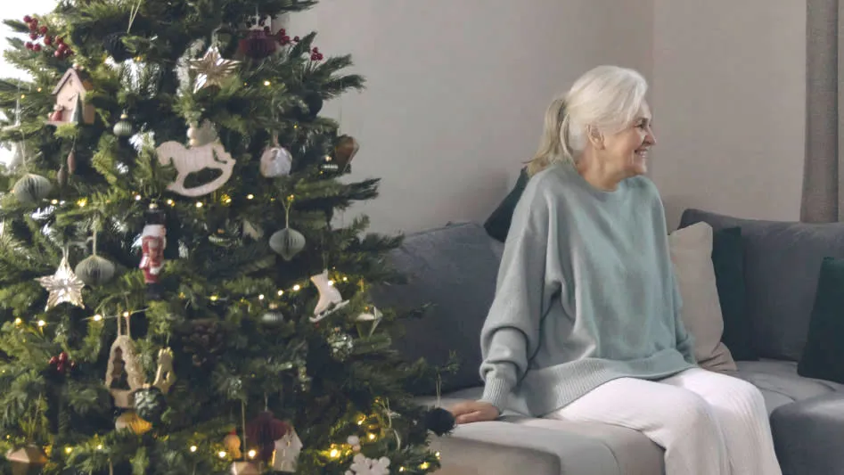An older woman sitting on a sofa beside a decorated holiday tree