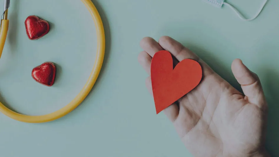 A hand holding a paper heart beside a stethoscope